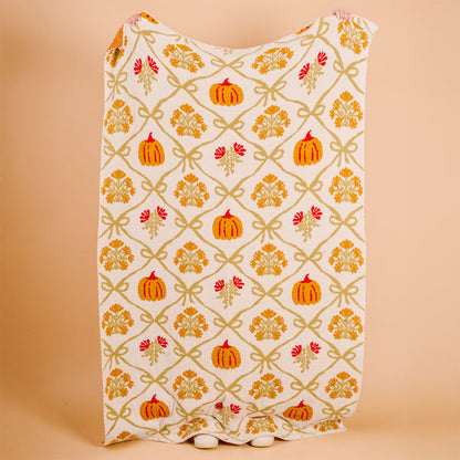 The Darling Effect's Patchwork Pumpkins Cozy Dreams Luxe Blanket, a white ultra-soft microfiber throw with orange pumpkins, yellow leaves, and green bows, is shown against a peach background with its bottom edge grazing the surface.