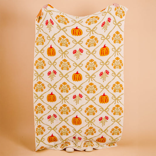 The Darling Effect's Patchwork Pumpkins Cozy Dreams Luxe Blanket, a white ultra-soft microfiber throw with orange pumpkins, yellow leaves, and green bows, is shown against a peach background with its bottom edge grazing the surface.
