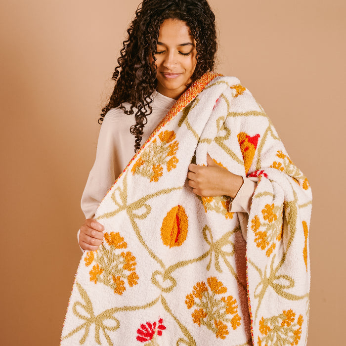 A young woman with curly dark hair smiles as she holds The Darling Effect’s Patchwork Pumpkins Cozy Dreams Luxe Blanket, an ultra-soft throw with a fuzzy white texture and vibrant orange and green patchwork pumpkin pattern.