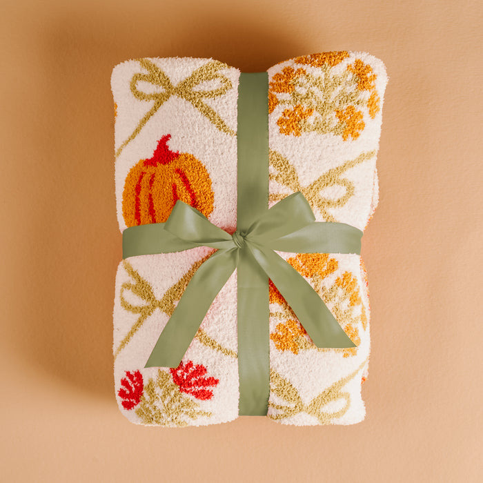The Darling Effect's Patchwork Pumpkins Cozy Dreams Luxe Blanket, ultra-soft and white with orange, red, and yellow pumpkin and leaf designs, is folded and tied with a green ribbon on a beige surface.