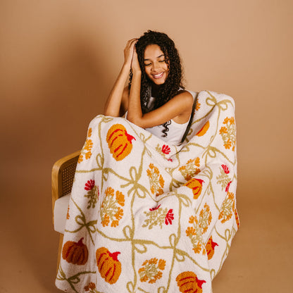 A woman with curly hair smiles while wrapped in The Darling Effect’s Patchwork Pumpkins Cozy Dreams Luxe Blanket—white with orange pumpkins, yellow leaves, and red flowers—against a beige background.