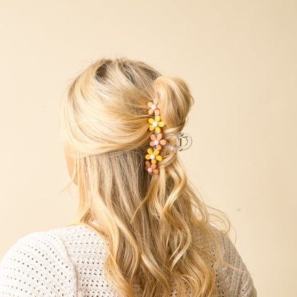 A woman with long blonde wavy hair, wearing a white textured top, is shown from behind. Her partial updo is secured with The Darling Effect’s Peachy Daisy Claw Clip—clear and adorned with pink and yellow daisy charms—against a beige background.