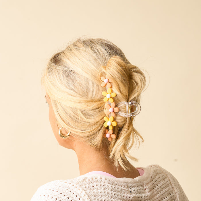 A woman with blonde hair styled in a loose updo, secured with The Darling Effect’s Peachy Daisy Claw Clip and pastel flower hair pins. She wears gold hoop earrings and a white knit top against a light beige background.
