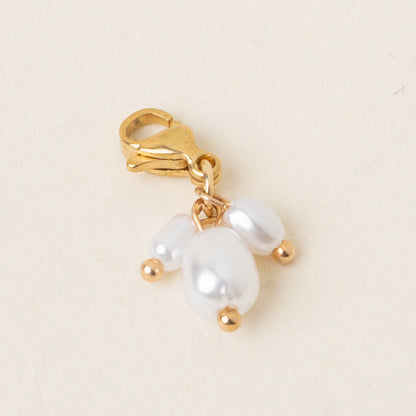 The Pearl Trio Jewelry Charm Gold by the-darling-effect features a gold clasp, three petite white pearls, and dainty gold beads. Shown on light cream, this charm is a sophisticated addition to your jewelry collection.