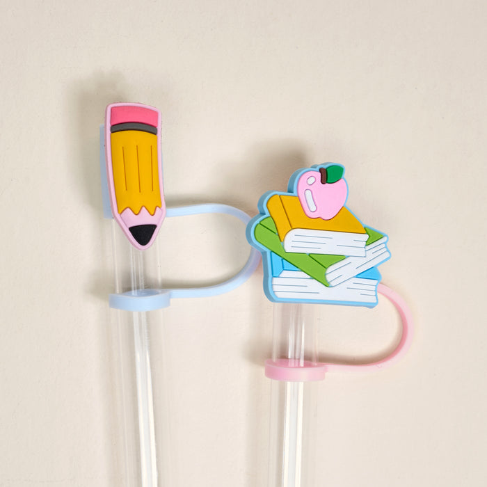 The Darling Effect's Pencil Me In Straw Topper Set includes two reusable clear straws with pastel loops and silicone toppers—a yellow pencil with pink eraser and a stack of books with an apple.