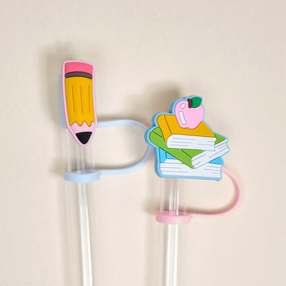 The Darling Effect's Pencil Me In Straw Topper Set includes two reusable clear straws with pastel loops and silicone toppers—a yellow pencil with pink eraser and a stack of books with an apple.