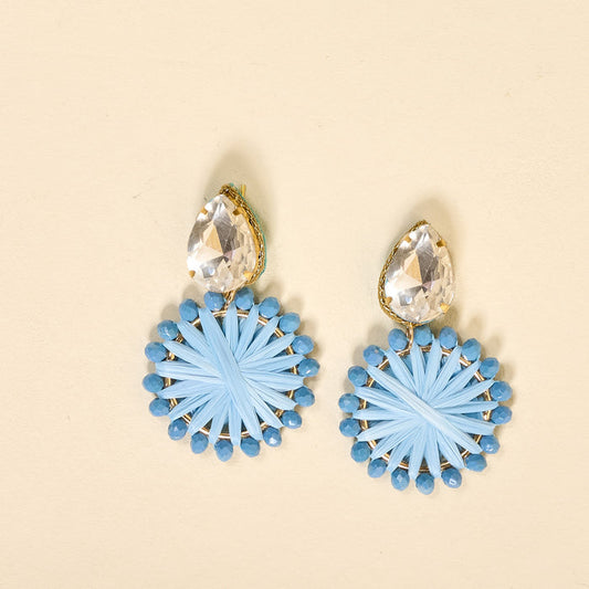 The Darling Effect's Periwinkle Radiant Gem Earrings feature large teardrop-shaped clear stones above circular, light blue woven designs edged with blue beads. Displayed on a light beige background.