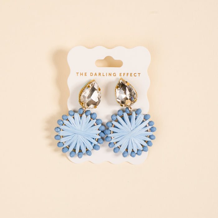 The Periwinkle Radiant Gem Earrings by The Darling Effect feature teardrop rhinestones and round, light blue woven accents. Displayed on a branded card against a beige background, they’re lightweight and perfect for everyday wear.