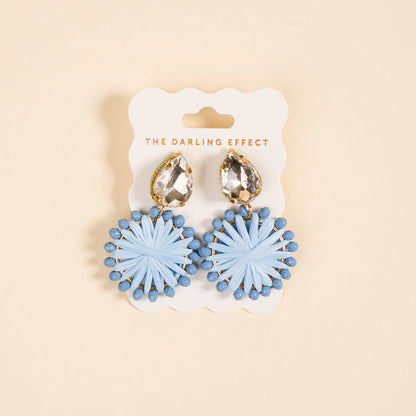The Periwinkle Radiant Gem Earrings by The Darling Effect feature teardrop rhinestones and round, light blue woven accents. Displayed on a branded card against a beige background, they’re lightweight and perfect for everyday wear.