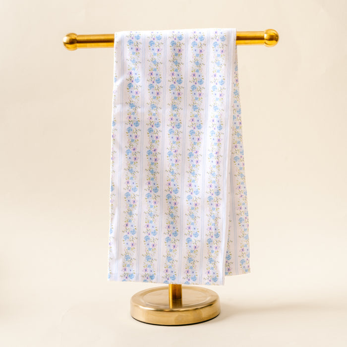 The Petal Parade Blue Tea Towel by the-darling-effect, featuring a blue and pink floral pattern on white cotton, is displayed neatly on a gold metal stand against a light beige background.