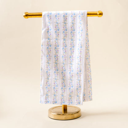 The Petal Parade Blue Tea Towel by the-darling-effect, featuring a blue and pink floral pattern on white cotton, is displayed neatly on a gold metal stand against a light beige background.