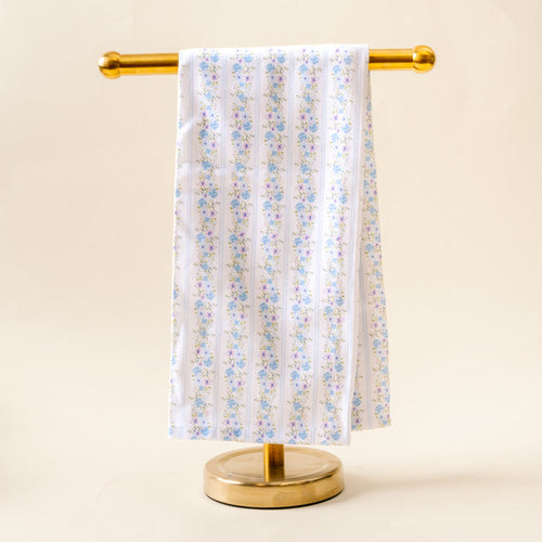 The Petal Parade Blue Tea Towel by the-darling-effect, featuring a blue and pink floral pattern on white cotton, is displayed neatly on a gold metal stand against a light beige background.
