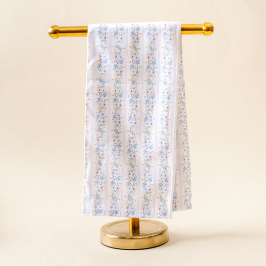 The Petal Parade Blue Tea Towel by the-darling-effect, featuring a blue and pink floral pattern on white cotton, is displayed neatly on a gold metal stand against a light beige background.
