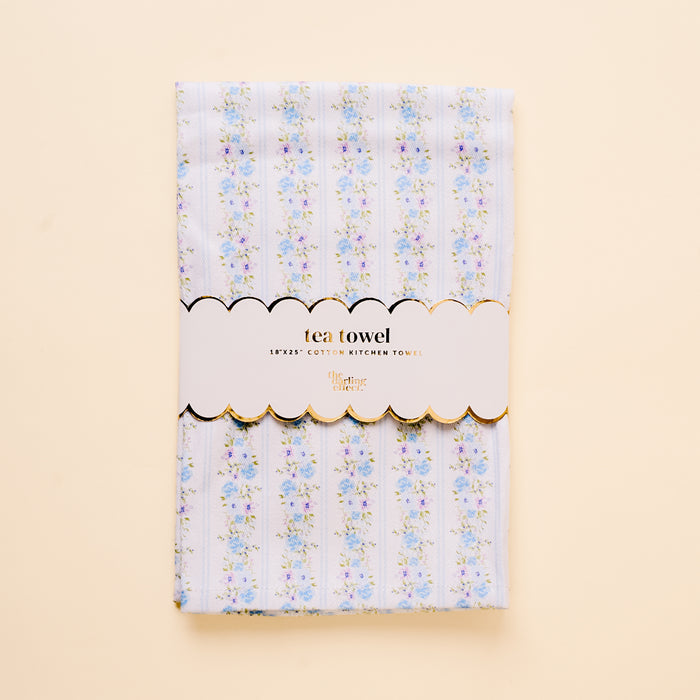The-darling-effect’s Petal Parade Blue Tea Towel is a cotton kitchen towel (18x25) with light blue and white stripes, blue and pink floral accents, and a white label trimmed in gold that reads "tea towel" and "18x25 COTTON KITCHEN TOWEL.