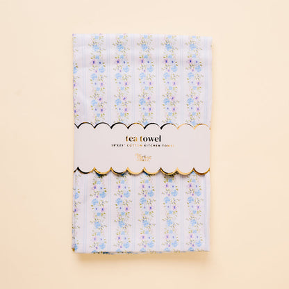 The-darling-effect’s Petal Parade Blue Tea Towel is a cotton kitchen towel (18x25) with light blue and white stripes, blue and pink floral accents, and a white label trimmed in gold that reads "tea towel" and "18x25 COTTON KITCHEN TOWEL.