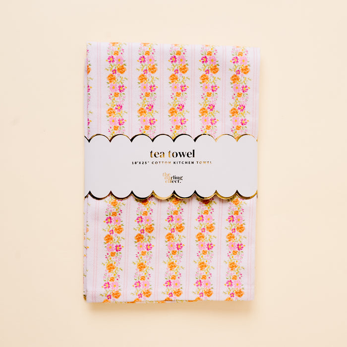 The Petal Parade Pink Tea Towel by the-darling-effect features a floral stripe pattern in pink, orange, and yellow. It’s folded with a white scalloped label reading "tea towel" and "100% cotton kitchen towel" on a light cream background.