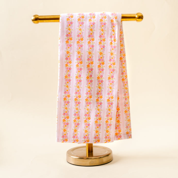 The Petal Parade Pink Tea Towel by the-darling-effect, featuring pink and orange florals, is draped over a gold stand against a light beige background.