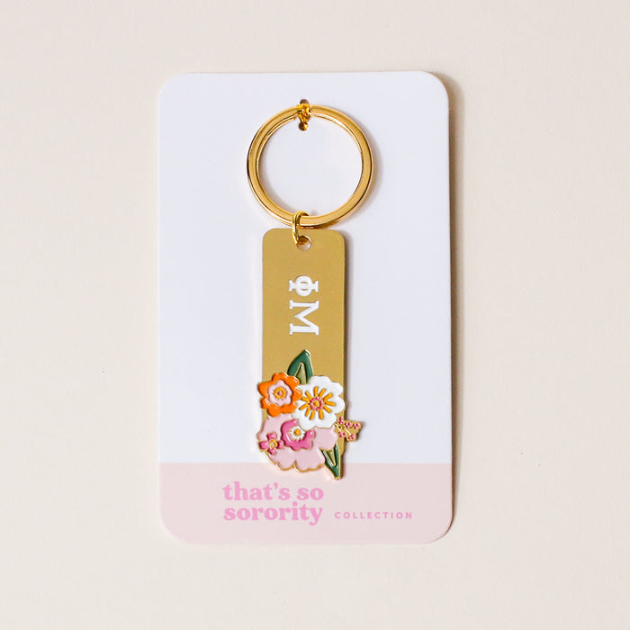The Darling Effect's Phi Mu Enamel Keychain features Greek letters with colorful florals on gold, attached to a white card with a pink edge reading "thats so sorority COLLECTION"—an ideal gift for any Phi Mu member.