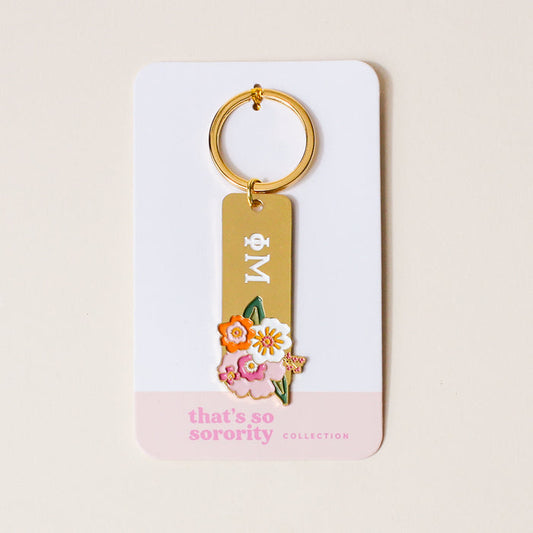 The Darling Effect's Phi Mu Enamel Keychain features Greek letters with colorful florals on gold, attached to a white card with a pink edge reading "thats so sorority COLLECTION"—an ideal gift for any Phi Mu member.