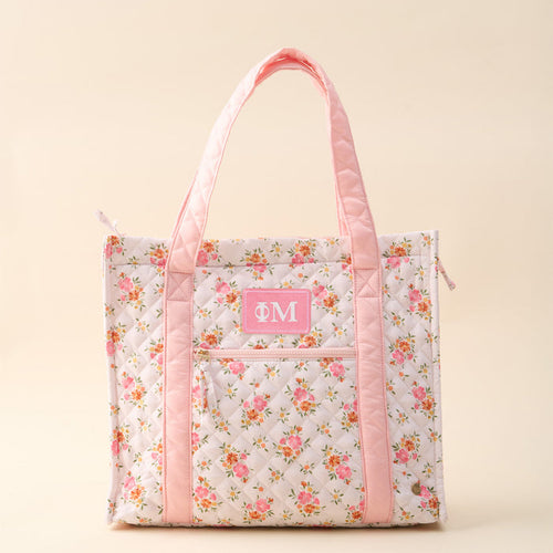 The Darling Effect’s Phi Mu The Everyday Quilted Tote Bag features a floral pattern, pink handles and trim, front zipper pocket, and a pink patch with white Greek letters—perfect for stylish everyday use.