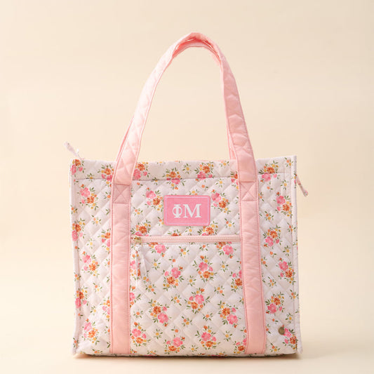 The Darling Effect’s Phi Mu The Everyday Quilted Tote Bag features a floral pattern, pink handles and trim, front zipper pocket, and a pink patch with white Greek letters—perfect for stylish everyday use.