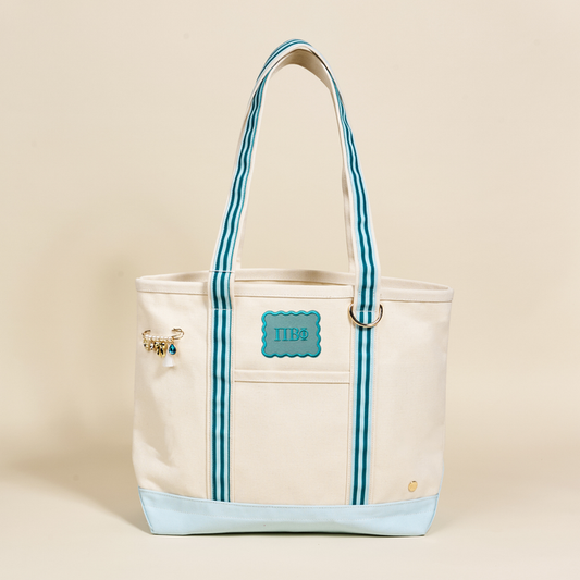 The Darling Effect's Pi Beta Phi Canvas Tote Bag features a beige canvas body, turquoise and white striped handles, monogrammed sorority letters, a beaded charm on one strap, a turquoise patch accent, and a light blue base—the ultimate sorority accessory.