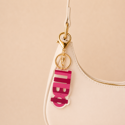 A close-up of a white handbag with a gold clasp and The Darling Effect’s Pi Beta Phi Stripes Acrylic Keychain in pink and white, featuring bold Cyrillic letters, attached to the handle against a plain light beige background.