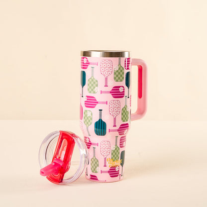 The-darling-effect Pickleball Party 40 oz Lifestyle Flip Straw Tumbler is tall and pink with a handle and colorful pickleball paddle pattern. A clear, leak-resistant lid with a red slider rests in front on a light surface against a beige background.