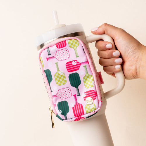 A hand holds a large white tumbler with a handle and straw, featuring the-darling-effect’s Pickleball Party Tumbler Fanny Pack—a water-resistant accessory with colorful pickleball paddle patterns on pink. Nails are painted pale pink.