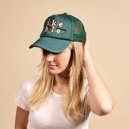 A woman models The Darling Effect's Pickleballer Trucker Hat—green with colorful "Pickleballer" lettering—paired with a white tee, touching the hat's brim against a plain beige background.