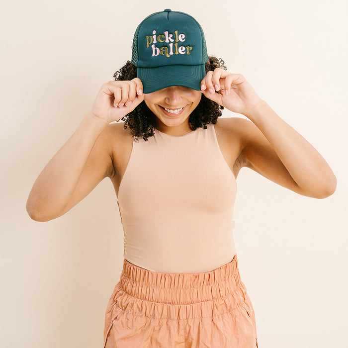 A smiling person wears The Darling Effect's Pickleballer Trucker Hat, a green embroidered cap, with both hands on the brim. She pairs it with a sleeveless beige top and light orange pants.