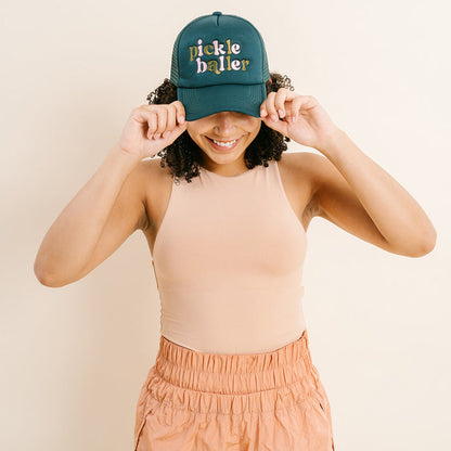 A smiling person wears The Darling Effect's Pickleballer Trucker Hat, a green embroidered cap, with both hands on the brim. She pairs it with a sleeveless beige top and light orange pants.