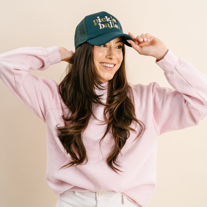 A woman with long brown hair wears a pink turtleneck sweatshirt and The Darling Effect’s Pickleballer Trucker Hat in green, featuring “Pickleballer” in gold and pink lettering.