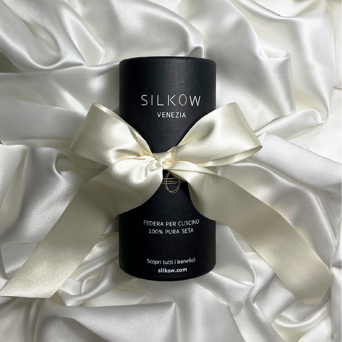 A black cylindrical pillowcase with the brand name SILKOW VENEZIA printed on it, presented on a white satin background with a white ribbon around it showcasing the  Pillowcase S22 100% Pure Silk Satin With Narrow Edge