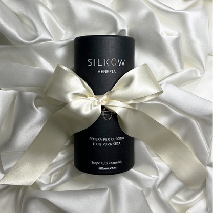 A black cylindrical pillowcase with the brand name SILKOW VENEZIA printed on it, presented on a white satin background with a white ribbon around it showcasing the  Pillowcase S22 100% Pure Silk Satin With Narrow Edge