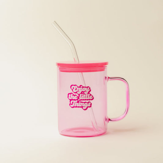 The Darling Effect’s Pink Enjoy the Little Things Color Glass Mug features a transparent pink body, handle, lid, and clear straw with retro white and pink text. This vibrant cup pops against a light background.