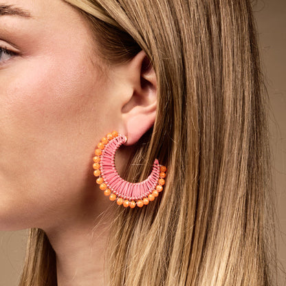 A close-up shows a woman wearing The Darling Effect's Pink Sunset It-Girl Earrings—large, pink, crescent-shaped and lightweight with orange bead accents. Her straight blonde hair is partly visible against a neutral background, perfect for everyday style.