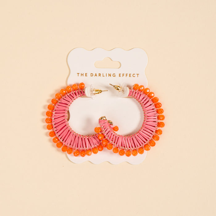 The Darling Effect’s Pink Sunset It-Girl Earrings feature lightweight pink wrapped material and bright orange beads, displayed on a branded white card against a beige background—perfect for everyday wear.