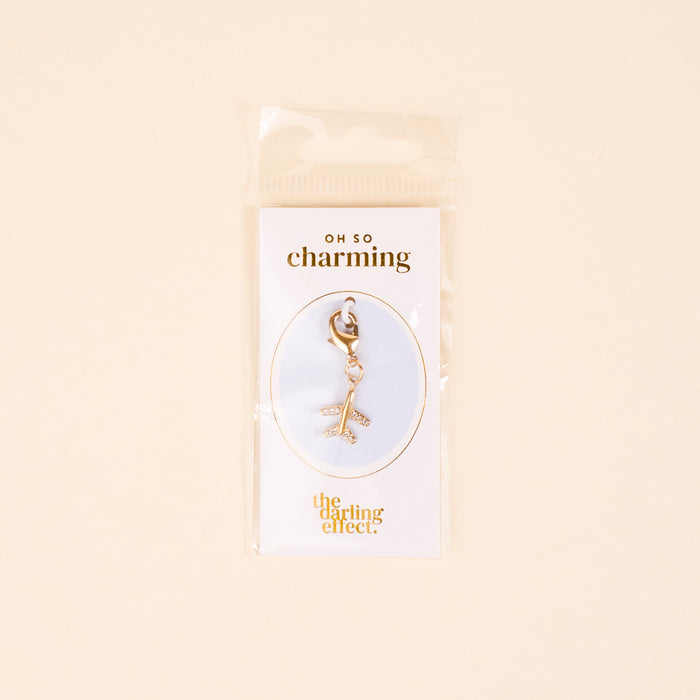 The Plane Jewelry Charm Gold by the-darling-effect features a gold airplane on a lobster clasp, displayed on a white card with gold "oh so charming" and "the darling effect" text, and comes packaged in a clear plastic bag.