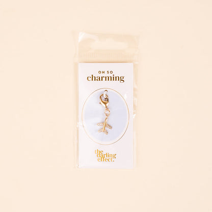 The Plane Jewelry Charm Gold by the-darling-effect features a gold airplane on a lobster clasp, displayed on a white card with gold "oh so charming" and "the darling effect" text, and comes packaged in a clear plastic bag.