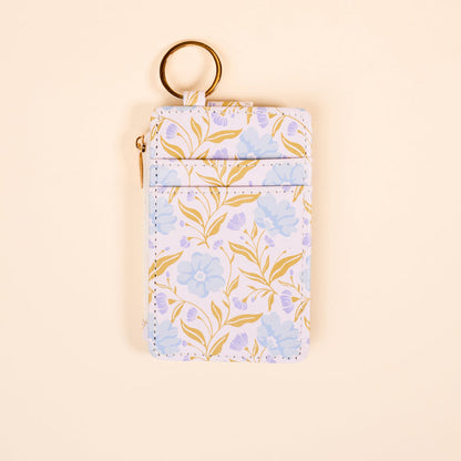 The Darling Effect’s Poppin' Petals Aqua Keychain Wallet, a slim card holder with blue flowers, yellow leaves, gold keyring, and zippered pocket, is displayed on beige. This stylish wallet is both functional and fashionable.