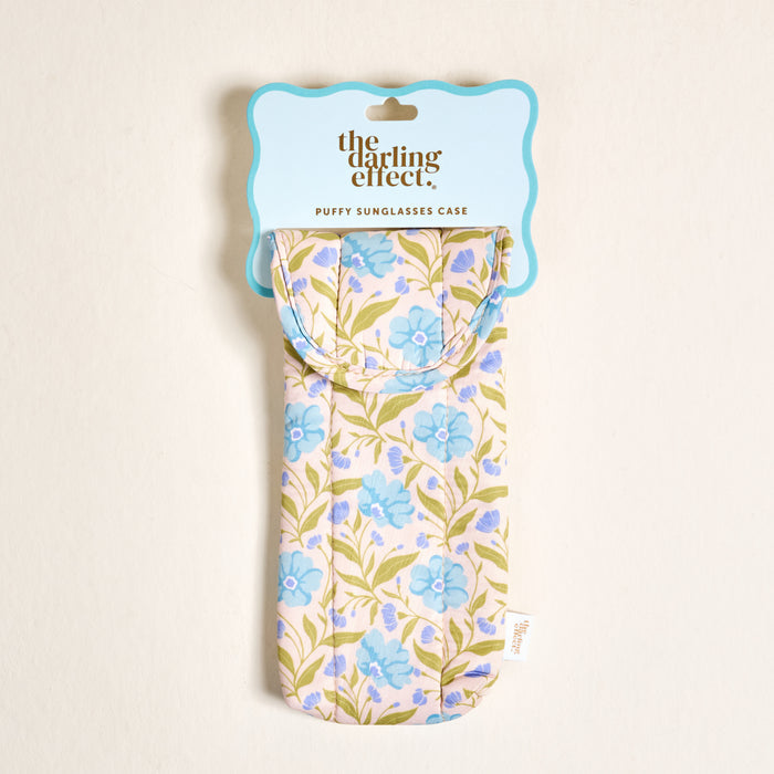 The Darling Effect Poppin' Petals Aqua Puffy Sunglasses Case features a pastel pink background with blue and green floral design, a magnetic flap closure, and hangs from a wavy light blue tag reading "the darling effect.