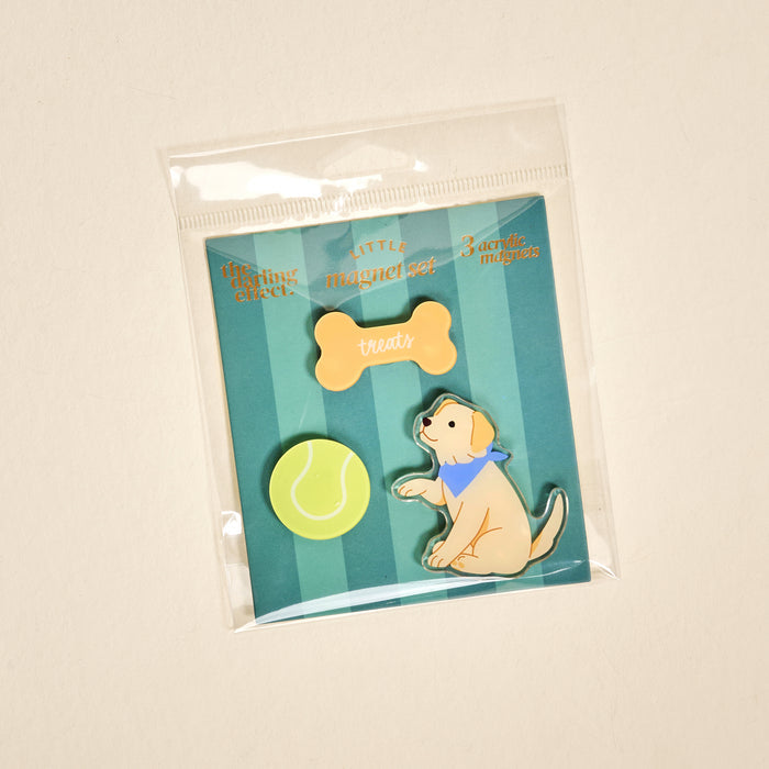 The Darling Effect's Puppy Playtime Little Magnet Set features three acrylic magnets—a yellow tennis ball, bone labeled “treats,” and a sitting yellow puppy in a blue bandana—perfect as charming fridge magnets or decorative accents.