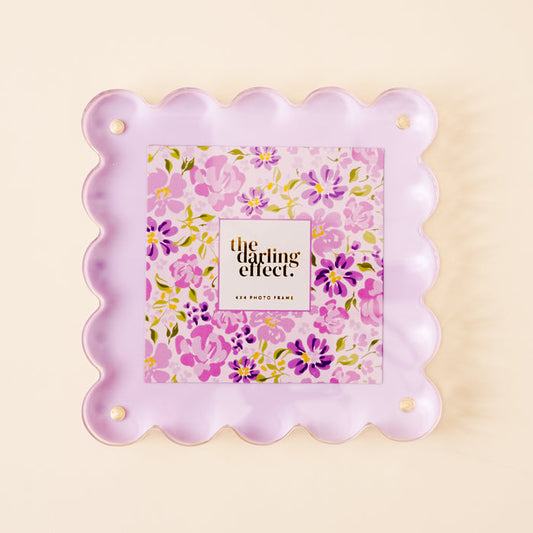 The Purple Square Scalloped Acrylic Frame by the-darling-effect features scalloped edges, pastel purple acrylic with magnetized panels, and encloses a floral print and “the darling effect” text. Holds a 4x4 photo in gold/black lettering.