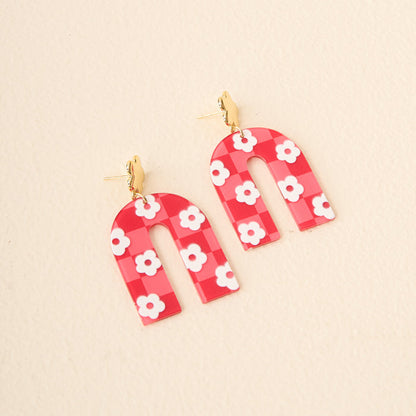 Show your school spirit with The Darling Effect's Red Game Day Acrylic Earrings—arch-shaped with a red and pink checkered pattern, white daisy accents, and gold studs—perfect for game day style.