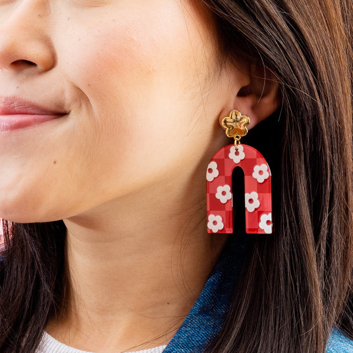 A close-up of a person wearing The Darling Effect’s Red Game Day Acrylic Earrings—bold, arch-shaped earrings with red and white floral patterns and gold flower studs—perfect for showcasing your school spirit on game day.