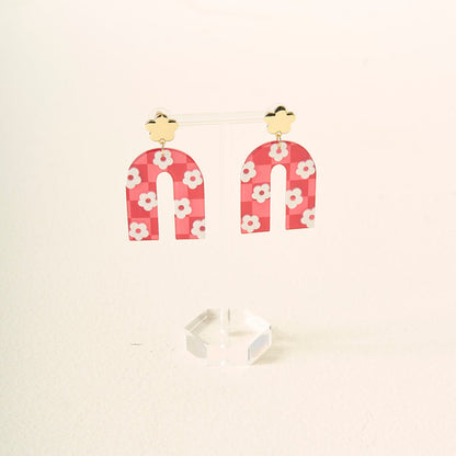 A pair of red, arch-shaped acrylic earrings with white flower patterns hang from gold-colored flower-shaped studs, displayed on a clear stand—Red Game Day Acrylic Earrings by The Darling Effect, perfect for showing your school spirit on game day.