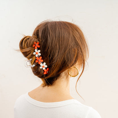 A woman wears her reddish-brown hair in a bun, secured with The Darling Effect’s Red Game Day Daisy Claw Clip featuring white and orange flowers—perfect for showing school pride. She pairs it with a white shirt and gold hoop earrings.