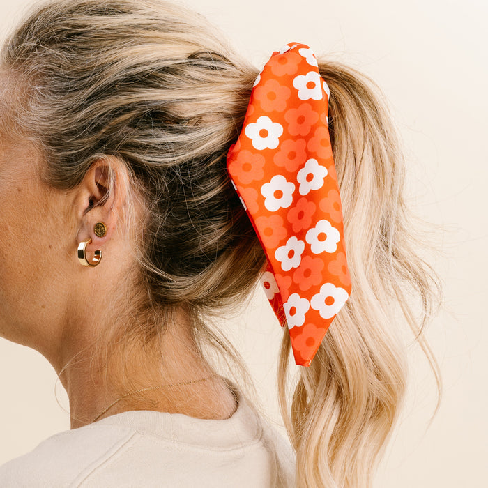 A person with long, blonde hair wears a ponytail tied with The Darling Effect’s Red Game Day Hair Scarf with Scrunchie—perfect for showing team spirit. They also wear gold earrings and a cream top against a plain, light background.