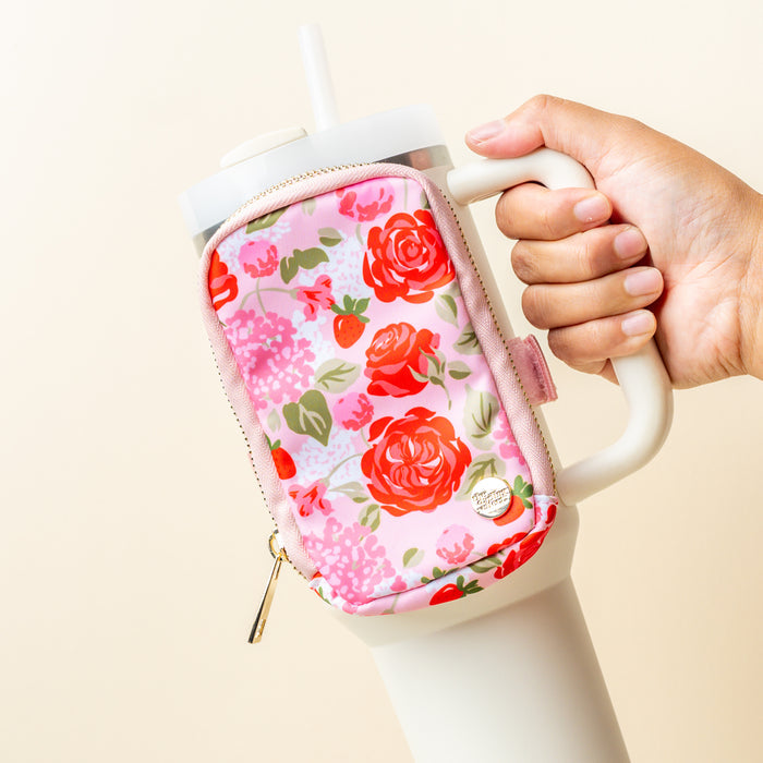 A hand holds a large white tumbler with a straw, accessorized by The Darling Effect’s Rose Garden Tumbler Fanny Pack—a pink pouch with a red and green floral print and gold zipper. The background is a plain, light beige color.